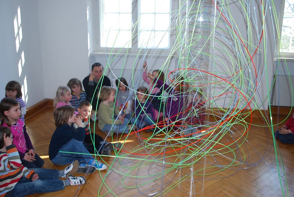 Eine Gruppe Kinder sitzt um ein Kunstwerk aus bunten Plastikstäben versammelt.