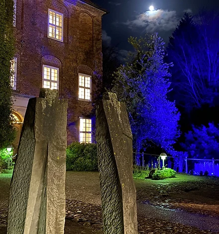 Zwei große, grob menschlich aussehende Steinskulpturen. Dahinter sieht man die Schlossfront mit erleuchteten Fenstern und blau angestrahlten Bäumen.