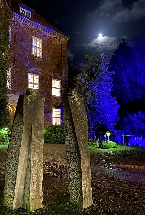 Zwei große, grob menschlich aussehende Steinskulpturen. Dahinter sieht man die Schlossfront mit erleuchteten Fenstern und blau angestrahlten Bäumen.