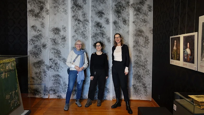 Neues Kunstwerk im Witwenzimmer Drei Frauen posieren vor einem Kunstwerk, Siebdruck-Bahnen von der Decke bis zum Boden eines Museumsraums.