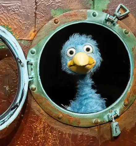 Eine Vogel-Puppe schaut aus einem Bullauge
