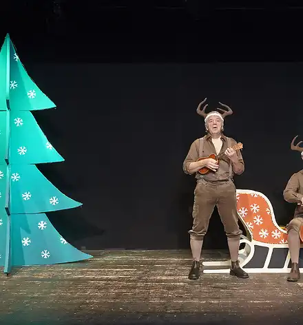 Links ein Tannenbaum, rechts zwei als Rentiere verkleidete Musiker mti deiner Ukulele und einem Cello.
