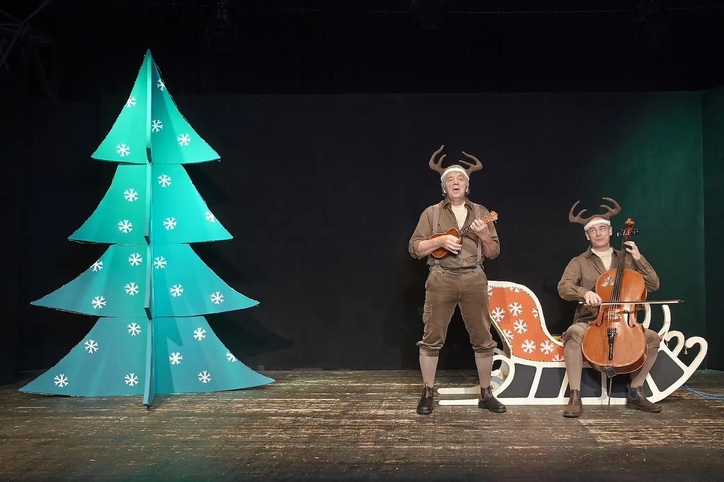 Links ein Tannenbaum, rechts zwei als Rentiere verkleidete Musiker mti deiner Ukulele und einem Cello.