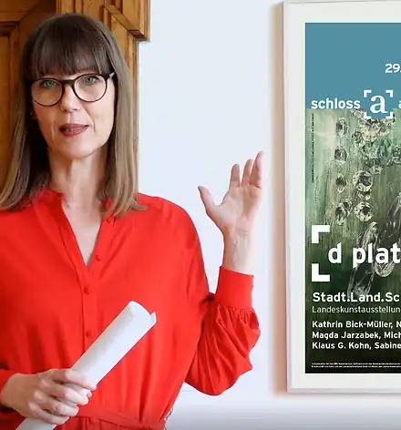 Art & Aperitif Claudia Rastzar zeigt auf das Plakat zur Ausstellung.