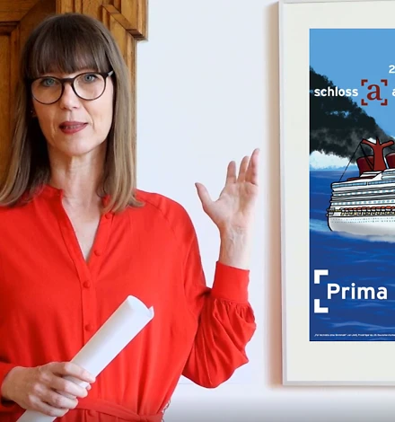 Kunstvermittlerin Claudia Rasztar trägt ein rotes Kleid und zeigt auf das Plakat zur Ausstellung.