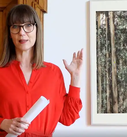 Claudia Rasztar zeigt auf ein Gemälde mit Bäumen.