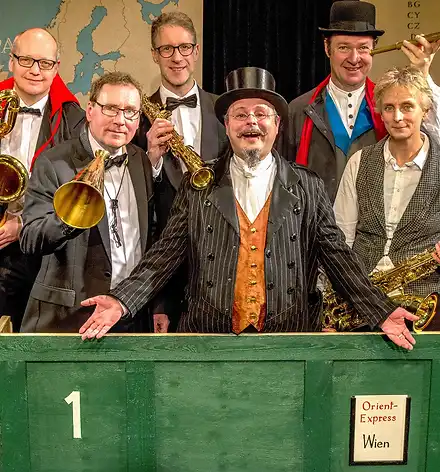 Die vier Musiker und zwei Schauspieler posieren in einer grünen Kiste mit der Aufschrift: 