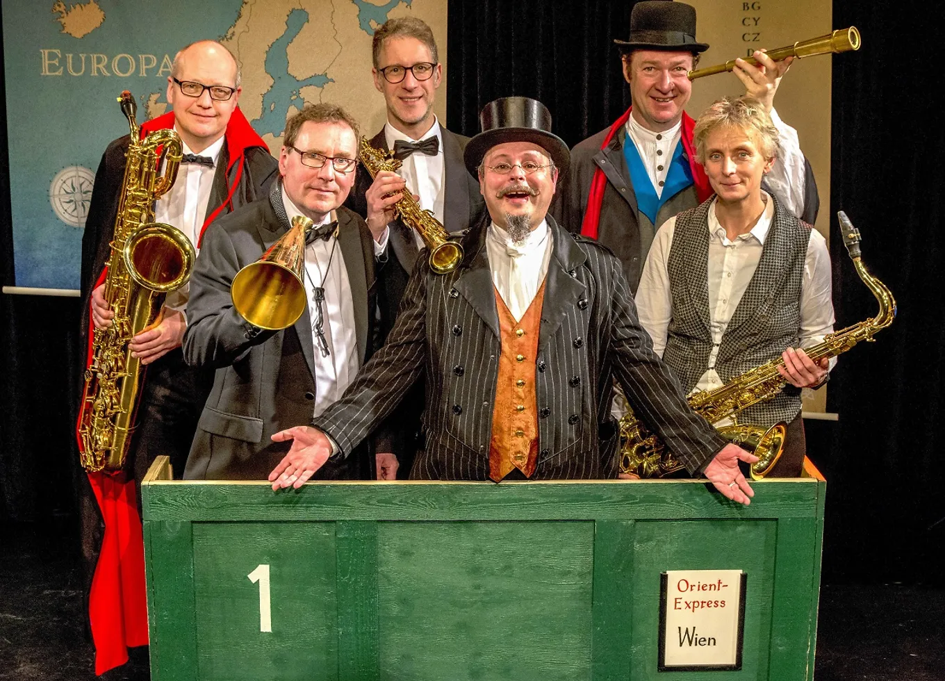 Die vier Musiker und zwei Schauspieler posieren in einer grünen Kiste mit der Aufschrift: 