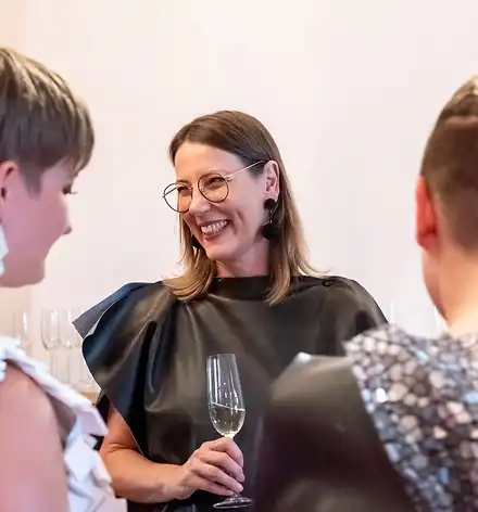 Ausstellungseröffnung Kuratorin Claudia Rasztar mit einem Sektglas im Gespräch mit zwei schick gekleideten jungen Frauen.