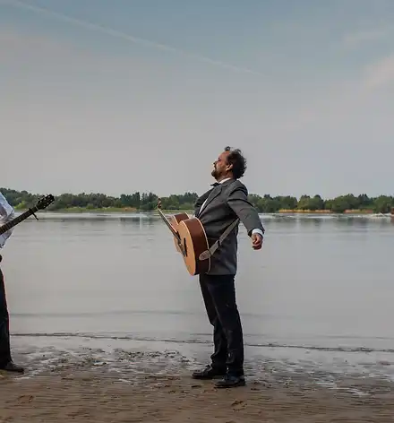 Foto von zwei Männern mit Gitarre vor der Elbe, auf der gerade ein Schiff vorbeifährt