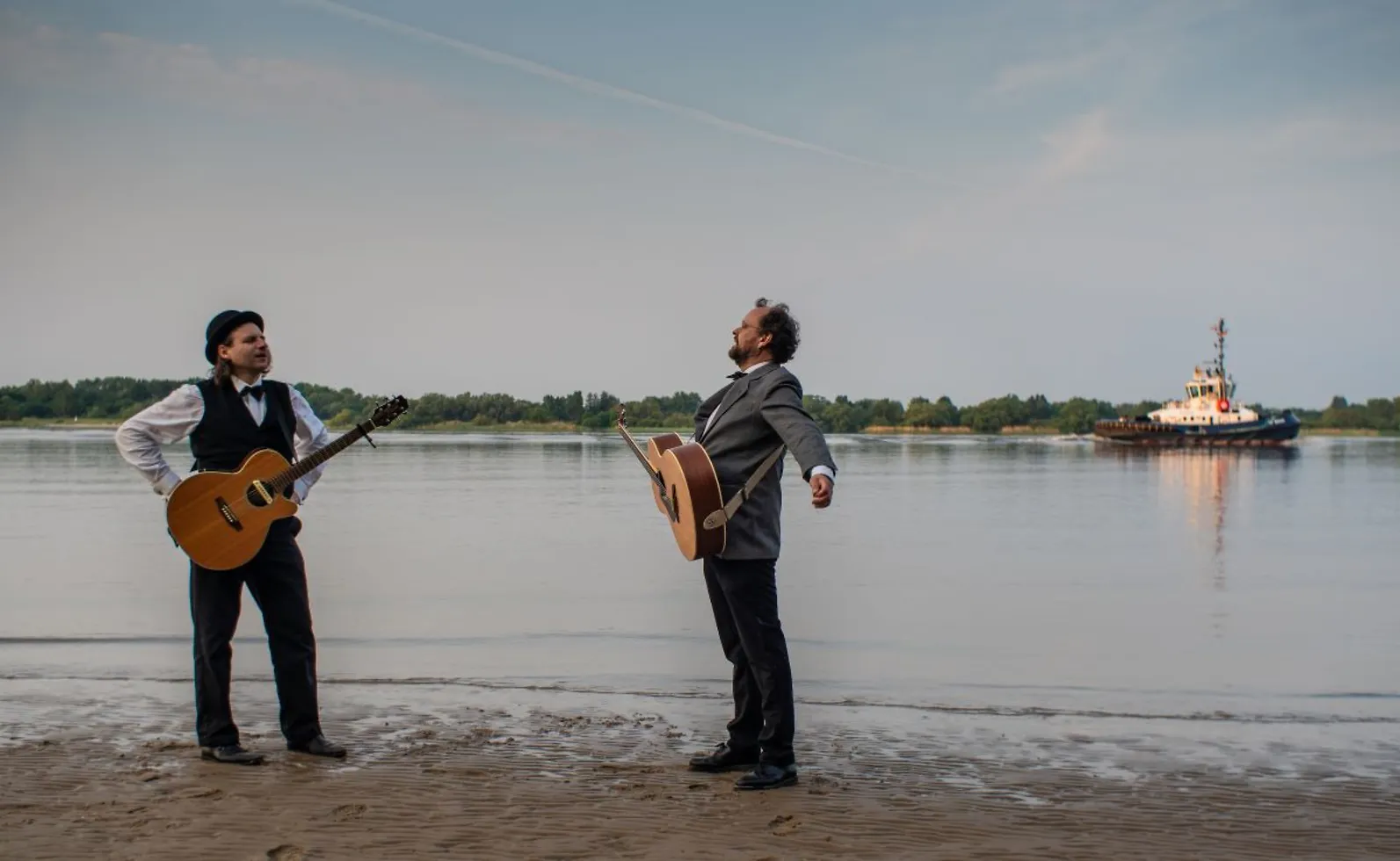 Foto von zwei Männern mit Gitarre vor der Elbe, auf der gerade ein Schiff vorbeifährt