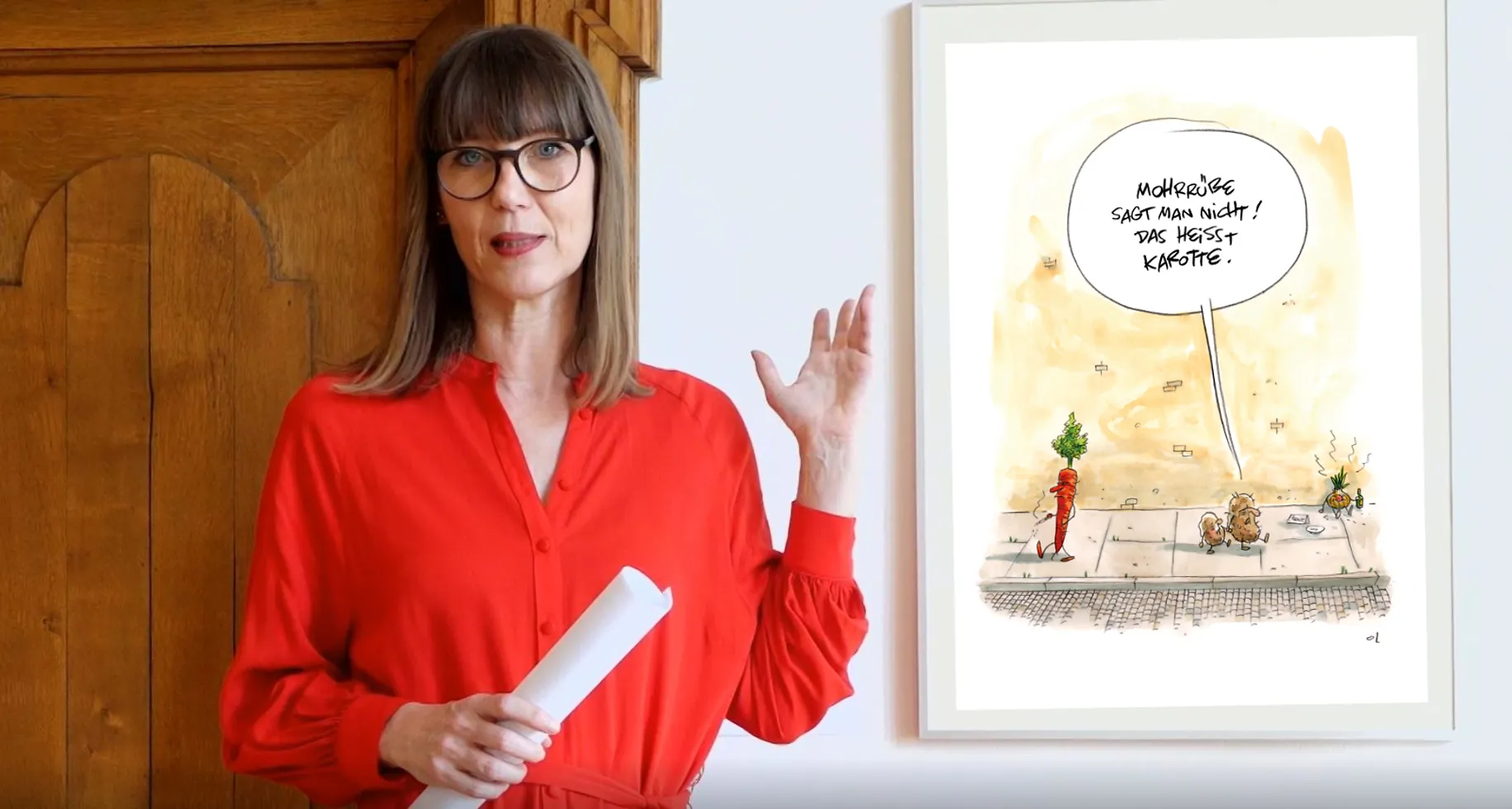 Claudia Rasztar zeigt auf eine gerahmte Karikatur.