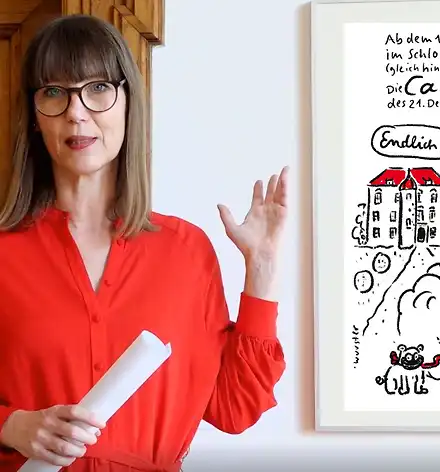 KarikaturKunst&Kaffee – Virtuell! Claudia Rasztar deutet auf eine gerahmte Karikatur.