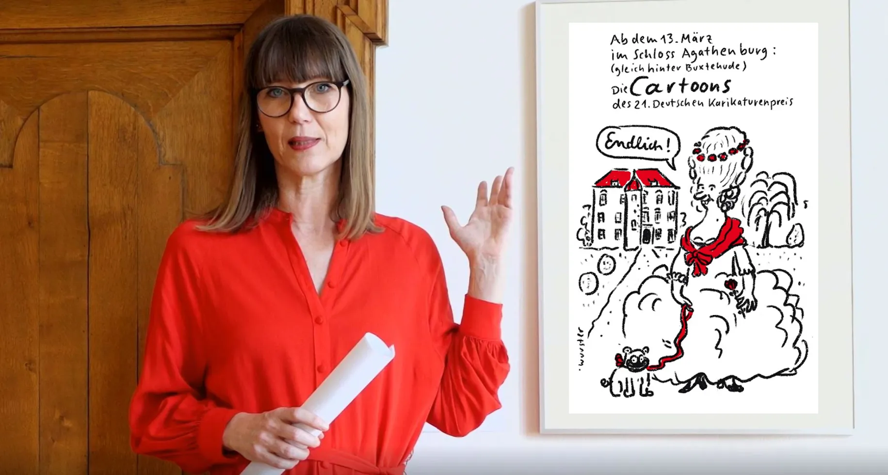 Claudia Rasztar deutet auf eine gerahmte Karikatur.