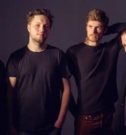 Foto der vier Musiker, alle in schwarz vor schwarzem Hintergrund