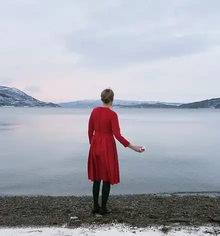 Eine Frau im roten Kleid vor arktischer Landschaft, mit einem Plastik-Joghurt-Becher in der Hand.