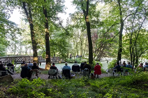 Am Rand des Teichs im Schlosspark sitzen Zuschauer und Blicken zum Saxophon Quartett auf der Insel hin. 